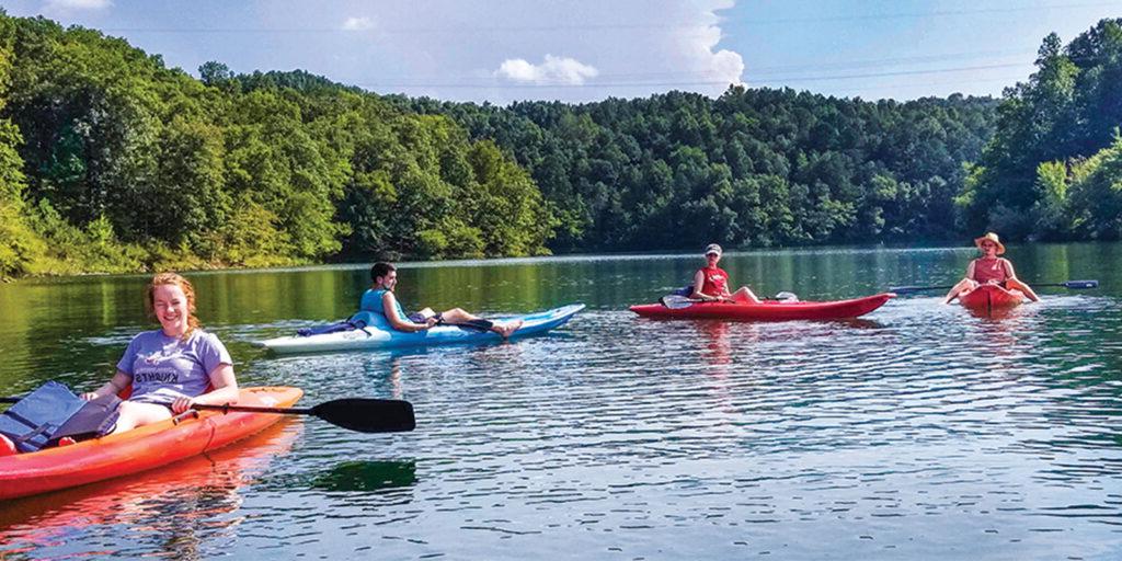 Four people paddle kayaks on a calm lake surrounded by lush, green trees on a sunny day.