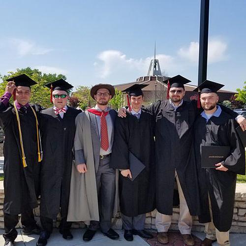 A group of six graduates wearing caps and gowns stand together outdoors, smiling at the camera. A prominent building with a spire is visible in the background under a clear sky.