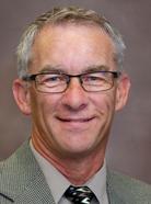 A man with short gray hair, glasses, and a greenish-gray blazer smiles at the camera against a plain background.